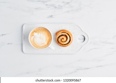 Freshly Baked Cinnamon Bun With Spices And Cocoa Filling And Coffee Or Cappuccino With Latte Art On White Serving Plate Over White Marble Background. Top View. Copy Space For Text. Swedish Breakfast.