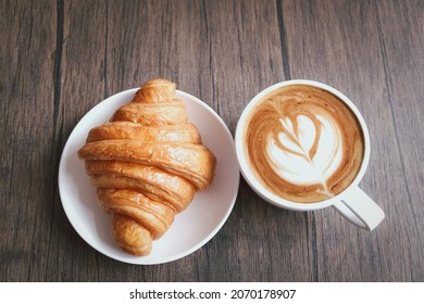 Freshly baked delicious croissant and cup of beautiful morning coffee on wooden breakfast table, top view