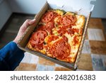 A freshly baked Detroit-style pizza, known for its thick crust and crispy cheese edges, loaded with tomatoes, mozzarella, and more, sits ready to eat in an open takeout box held by a male hand.

