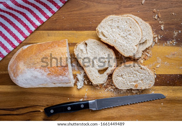 Freshly baked\
Italian Bread, sliced with a serrated bread knife on a wooden\
cutting board with a red and white\
towel