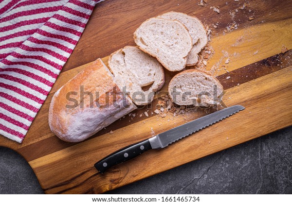 Freshly baked Italian Bread, sliced with a\
serrated bread knife on a wooden cutting board with a red and white\
towel on a slate\
countertop