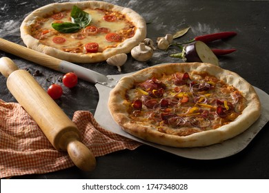 Freshly baked pizza margherita with cherry tomatoes and pizza with bacon, onion and pepper lie on a shovel among the ingredients in flour on a black table.