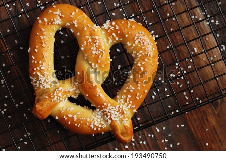 Freshly baked soft pretzel with generous sprinkling of coarse salt on wire cooling rack over rustic dark wood.  Closeup from above.