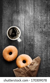 Freshly Baked Sugared Ring Doughnuts With A Mug Of Full Roast Filter Coffee And Brown Paper Packaging On A Grungy Old Wooden Table In A Rustic Kitchen For Breakfast Snack