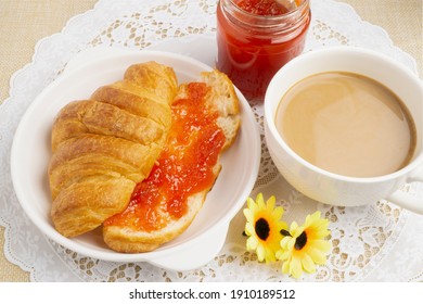 Freshly baked tasty croissant cut in half spreaded with strawberry jam served with hot coffee on white lace placemat