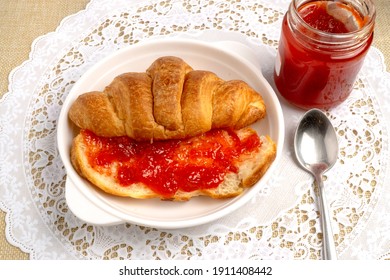 Freshly baked tasty croissant cut in half spreaded with red strawberry jam on white lace placemat