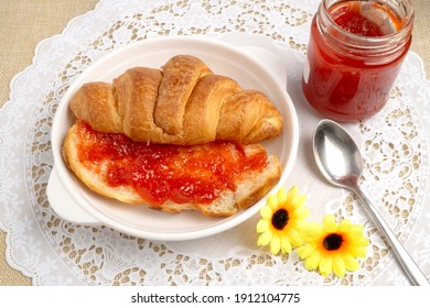 Freshly baked tasty croissant cut in half spreaded with red strawberry jam on white lace placemat