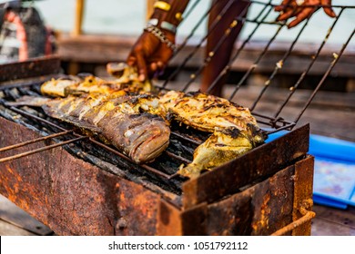 Freshly caught tropical ocean fish being grilled in a folded wire grill basket, outdoors. Hand of fisherman visible, checking doneness. Onboard a wooden fishing dhow. Lamu Island, Kenya, Africa.