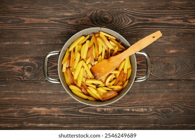 Freshly cut potato fries are frying in a large pot, their golden color starting to develop. A wooden spatula rests inside the pot, ready to stir the crispy fries on a rustic wooden table. - Powered by Shutterstock