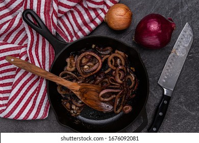 Freshly fried onions in a cast iron skillet, with a wooden spoon, Santoku knife, fresh yellow and red onions next to a red and white striped dish towel on a slate countertop - Powered by Shutterstock