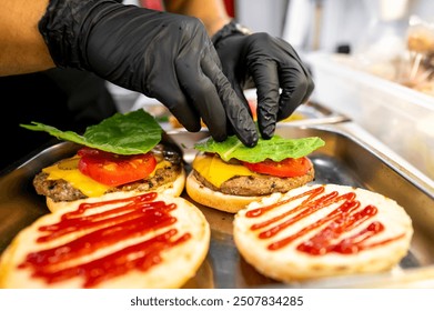 Freshly made burgers with cheese, lettuce, and tomato, ketchup on buns. Hands in gloves assembling meal. Perfect for fast food, culinary, and restaurant themes - Powered by Shutterstock