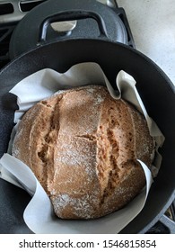 Freshly Made Loaf Of Round Bread In The Black Cast Iron Oven Pan With A Lid On The Side. Homemade Bread With Cuts On The Side Laying On A Parchment Paper.