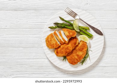 fried breaded chicken breasts with roast asparagus and lime on white plate on white wooden table, horizontal view from above, flat lay, copy space