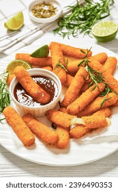 fried breaded mozzarella sticks served with dipping sauce, lime slices and fresh rosemary on white plate on white wooden table with ingredients, vertical view, close-up