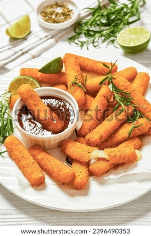 fried breaded mozzarella sticks served with dipping sauce, lime slices and fresh rosemary on white plate on white wooden table with ingredients, vertical view, close-up