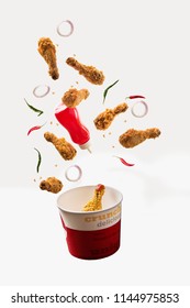 fried chicken legs flying with ketchup bottle, green and red chilli, onion rings and paper container over plain background
