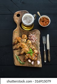 Fried chicken wings on rustic serving board, spicy tomato sauce, herbs and mug of light beer over black wooden backdrop, top view