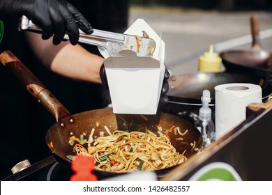 Fried chinese japanese noodles with vegetables and shrimps in takeaway box.Food delivery. Chef putting noodles in carton box to go from open kitchen. Asian street food festival