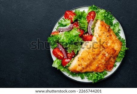 Fried cod fish with salad garnish from lettuce, cherry tomatoes and red onion with sesame seeds, black table background, top view
