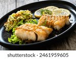 Fried codfish loin served with cabbage salad and lemon slices on black plate on wooden table 