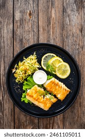 Fried codfish loin served with cabbage salad and lemon slices on black plate on wooden table 
