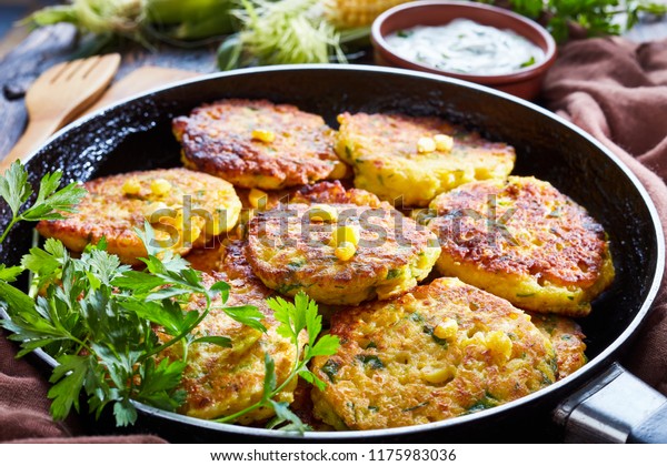 fried corn fritters in a skillet with yogurt\
dipping sauce and fresh sweet corn in cob on an old wooden kitchen\
table, close-up