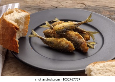 Fried Crunchy Little Fish On The Black Matte Plate. Stripe Textile Glass Of Water And Broke Up Bread. Wooden Background. Atlantic Horse Mackerel. 