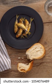 Fried Crunchy Little Fish On The Black Matte Plate. Stripe Textile Glass Of Water And Broke Up Bread. Wooden Background. Atlantic Horse Mackerel. 