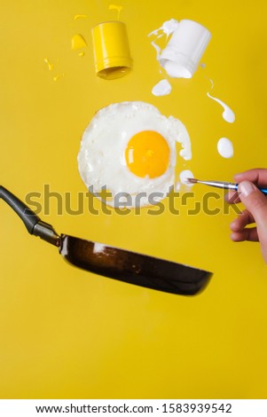 A fried egg and a pan with cans of paint and a brush rise into the air, and a hand with a brush is painted