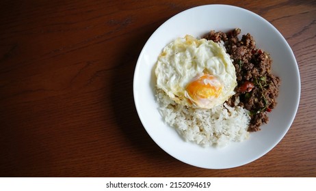 Fried egg and Thai basil beef or stir fried spicy minced beef with holy basil leaves served with jasmine rice in white ceramic plate on dark wooden table. Popular Thai dish “Pad Krapow Neua Kai Dao”.