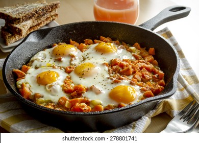 Fried eggs and sweet potato hash in cast iron skillet sitting on yellow striped kitchen towel with whole grain toast and grapefruit juice