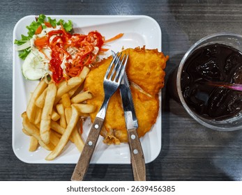 Fried fish with crispy fries and fresh salad, served with spicy sauce on a white plate, accompanied by iced tea on a dark wooden table.