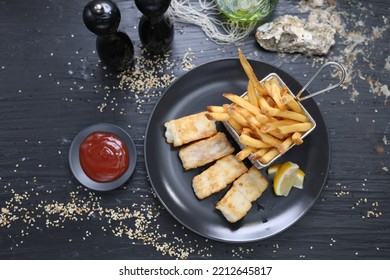 Fried Fish Fillets Served With Potato Fries In A Metal Serving Basket, On A Black Plate, Top View.