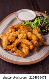 Fried Frog Legs With Salad, Arugula And Parsley Sauce On An Oval Plate And On A Wooden Background. Vertical Orientation