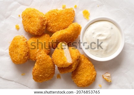 Fried gluten free cornflake crumb chicken nuggets next to a white ceramic bowl of white sauce on white baking paper. Top view.
