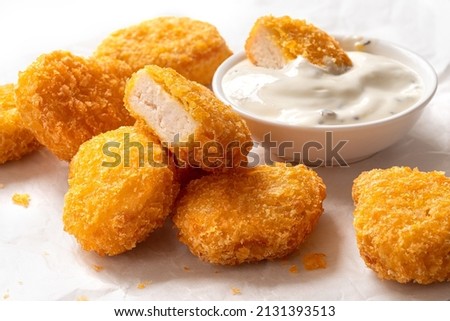 Fried gluten free cornflake crumb chicken nuggets next to a bowl of white sauce on white baking paper. Dipped nugget.
