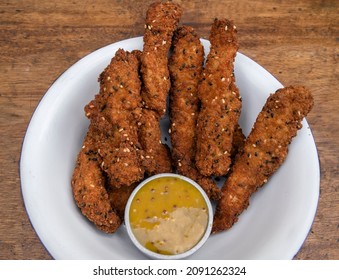 Fried Gourmet Food. Overhead View Of Deep Fried Chicken Sticks Breaded With Seeds And A Dip Made Of Mustard And Honey, In A White Bowl On The Table.