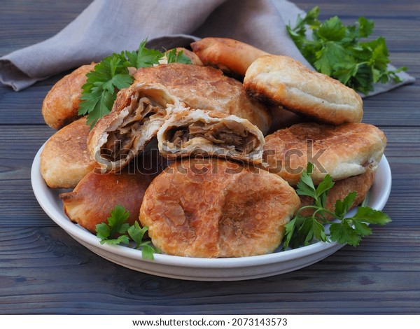 Fried pies with cabbage in a\
plate with a napkin, herb parsley on a dark wooden table, closeup.\
Traditional delicious hot snack pirozhki. Russian, Ukrainian\
cuisine