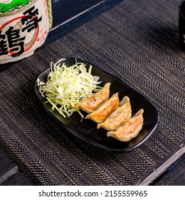 Fried pork gyoza dumplings with salad in the traditional Japanese ramen restaurant, with a black plate on a black mat and Japanese fonts sake barrel translation: Snow Crane
