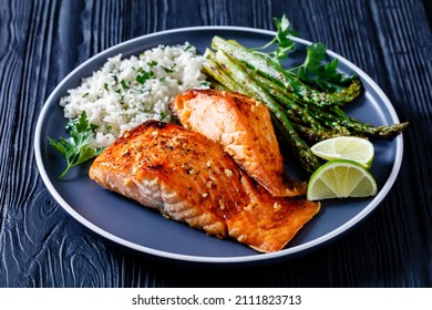 Fried Salmon Fish Fillets With Asparagus, Jasmine Rice And Lime On A Plate On A Black Wooden Table, Horizontal View From Above, Close-up