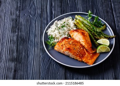 Fried Salmon Fish Fillets With Asparagus, Jasmine Rice And Lime On A Plate On A Black Wooden Table, Horizontal View From Above, Close-up