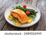 Fried salmon steak with cooked green asparagus, cherry tomatoes and lemon slices served on white plate on wooden table