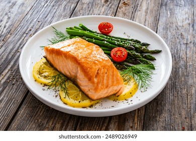 Fried salmon steak with cooked green asparagus, cherry tomatoes and lemon slices served on white plate on wooden table