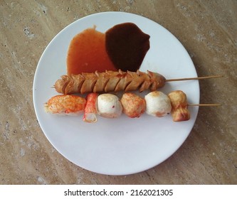 A fried sausage satay and a fried salmon ball, crab stick, chikuwa, crab claw, and fish ball satay on a plate with barbeque sauce and black pepper for dipping