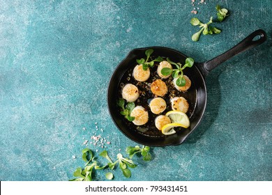 Fried scallops with butter lemon spicy sauce in cast-iron pan served with green salad over turquoise texture background. Top view, copy space
