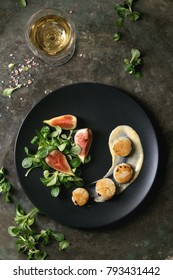 Fried Scallops With Lemon, Figs, Sauce And Green Salad Served On Black Plate With Glass Of White Wine Over Old Dark Metal Background. Top View, Space. Plating, Fine Dining