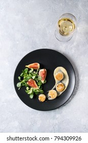 Fried Scallops With Lemon, Figs, Sauce And Green Salad Served On Ceramic Black Plate With Glass Of White Wine Over Gray Texture Background. Top View, Space. Plating, Fine Dining