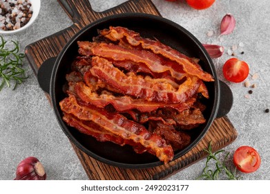 Fried slices of bacon for breakfast in a cast iron pan with tomatoes, garlic, rosemary and spices on a concrete background. Top view