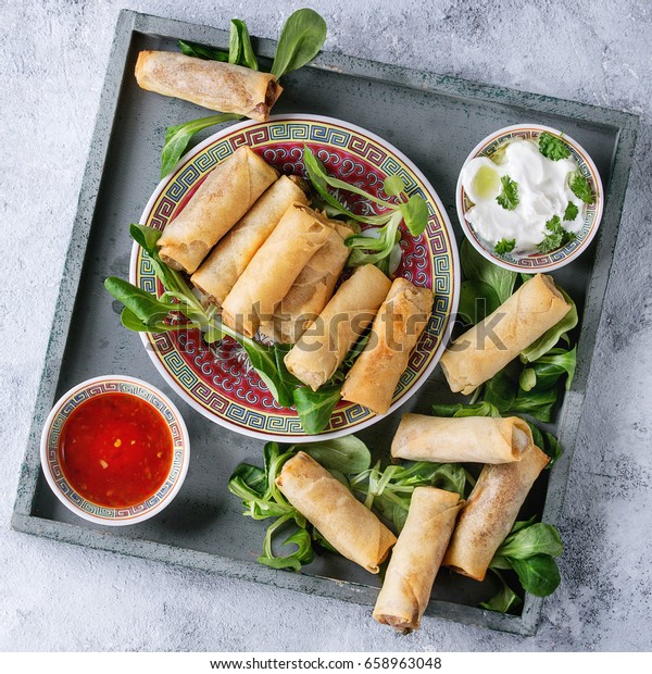 Fried spring rolls with red and white sauces,\
served in china plate on square wood tray with fresh green salad\
over gray blue texture background. Flat lay, space. Asian food.\
Square image