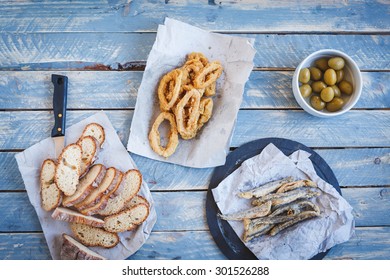 Fried Squid Rings Breaded On Paper With Fried Sardines, Marinated Cucumber And Sandwich Bread Baguette From Above On Kitchen Blue Wooden Table. Dinner And Seafood Concept. Rustic Style.